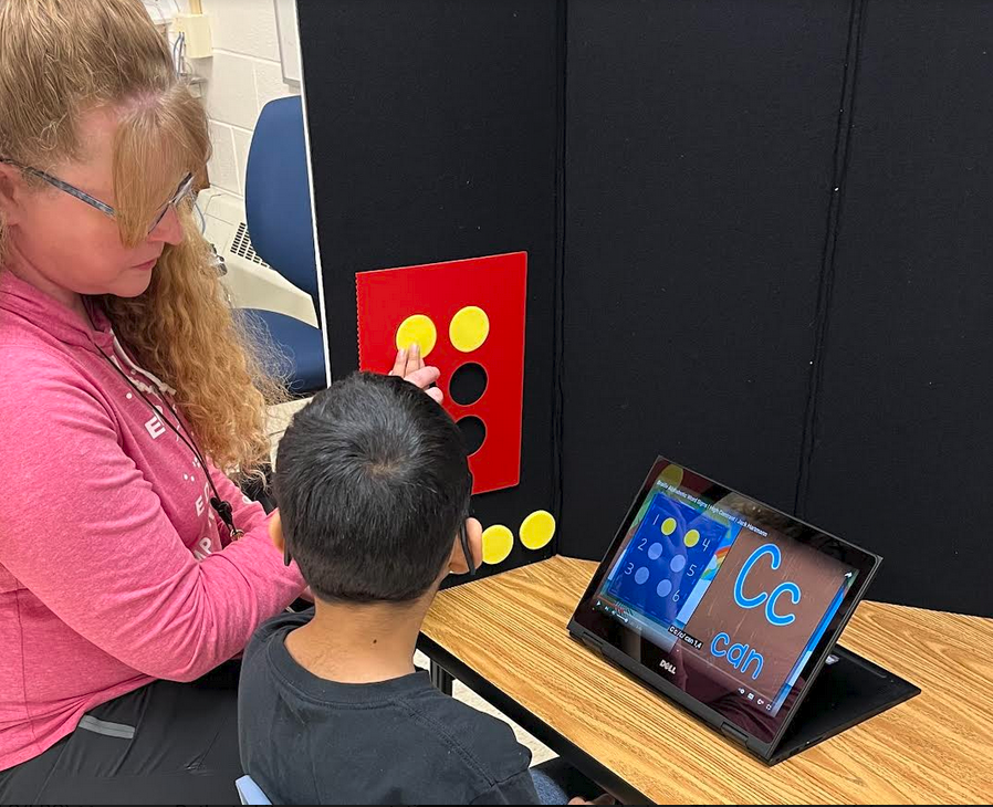 Teacher with young student learning the braille letter C with iPad screen showing video and manipulatives