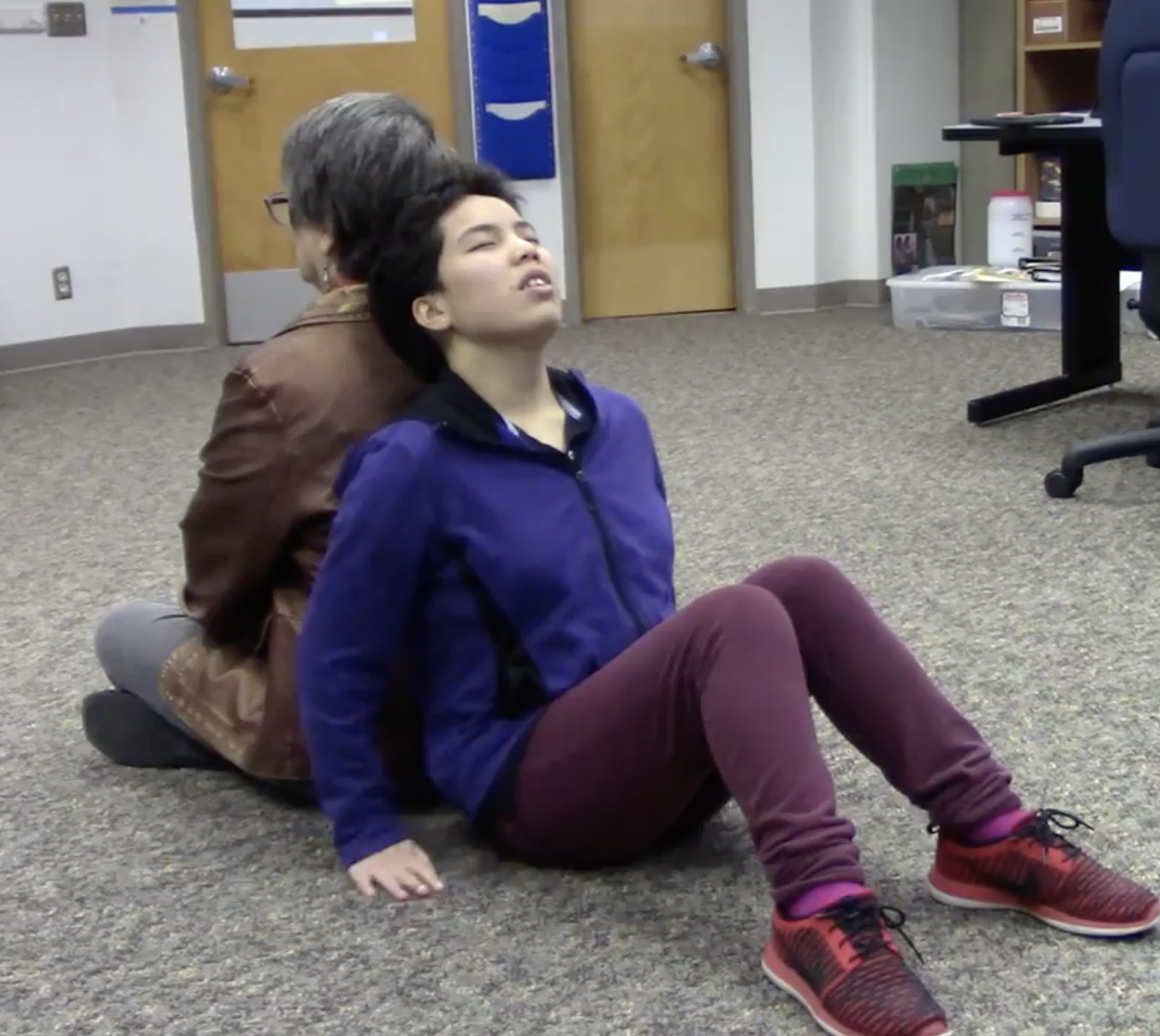 A woman and teenage girl sit back to back on a carpeted floor