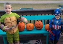 Two boys wearing Halloween costumes standing next to pumpkins on a bench