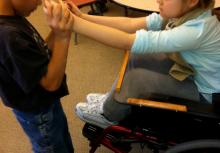 A boy holds the outstretched hands of a girl in a wheelchair.