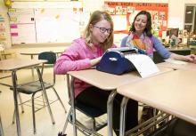 A girls with glasses uses a braille writer at a desk in a public school classroom.