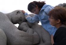 A teenage girl examines a stone statue of human figures as an adult looks on.