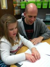 A girl reads a braille book while a man looks on.