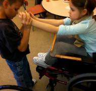 A boy holds the outstretched hands of a girl in a wheelchair.