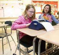 A girls with glasses uses a braille writer at a desk in a public school classroom.