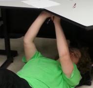 A student lies on the floor and writes on the underside of a desk above her.