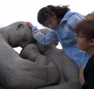 A teenage girl examines a stone statue of human figures as an adult looks on.