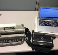  braillewriter, braille eraser, and a braille notetaker paired with a computer.  The braille display reads “I like to bake”.”