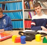 Two boys read  braille textbooks with math manipulatives on the table.
