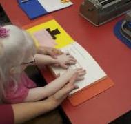Young girl with glasses reads a braille book with adult guidance.