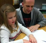 A girl reads a braille book while a man looks on.