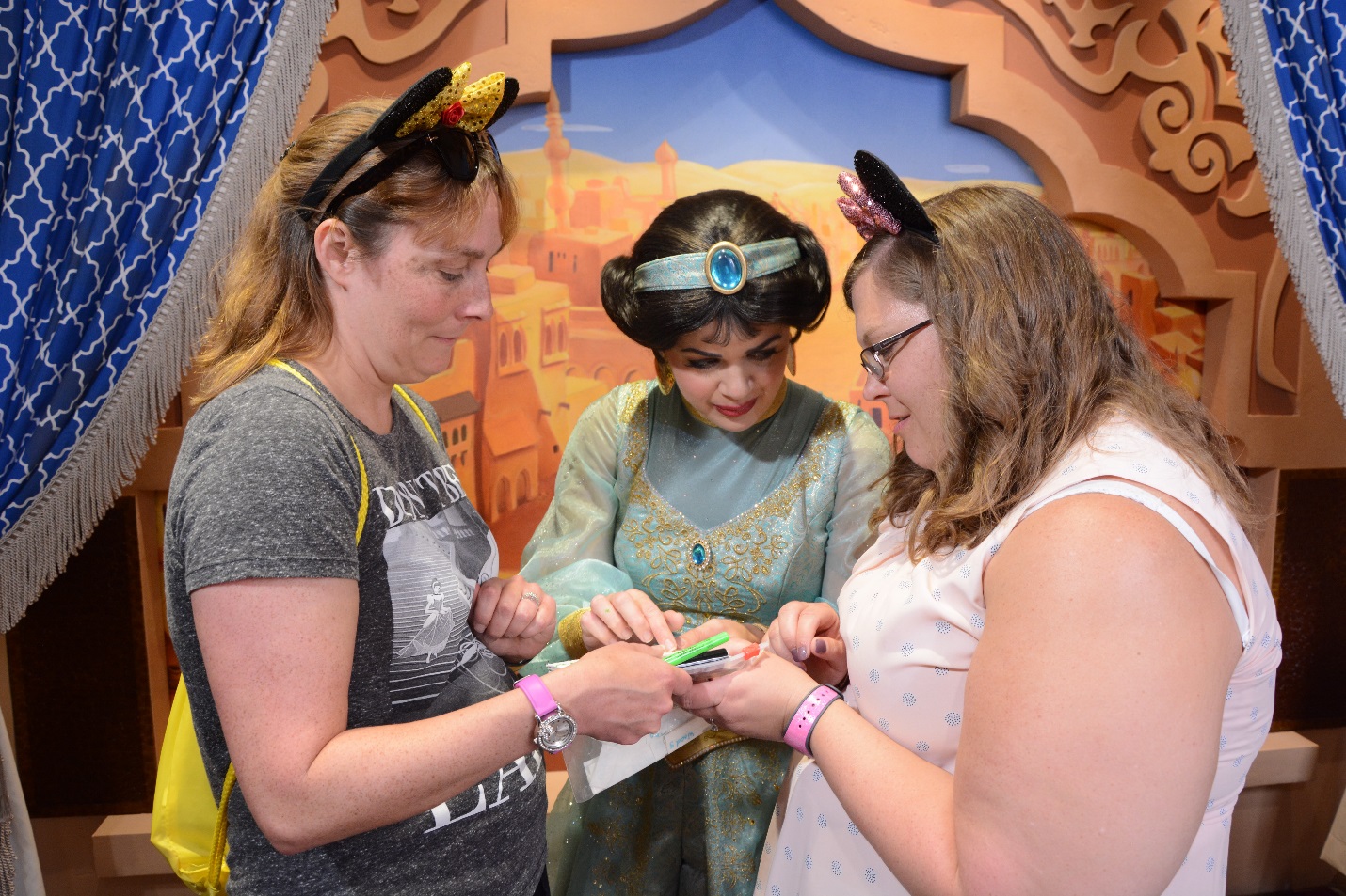 a woman, a young woman, and a woman dressed as Jasmine from Aladdin stand together while Jasmine signs the autograph book