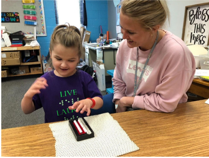 Young girl smiling working with a Beginner’s Abacus with a paraprofessional/braille transcriptionist. She is wearing a print/braille shirt with “Live” in print and “well” in braille visible]