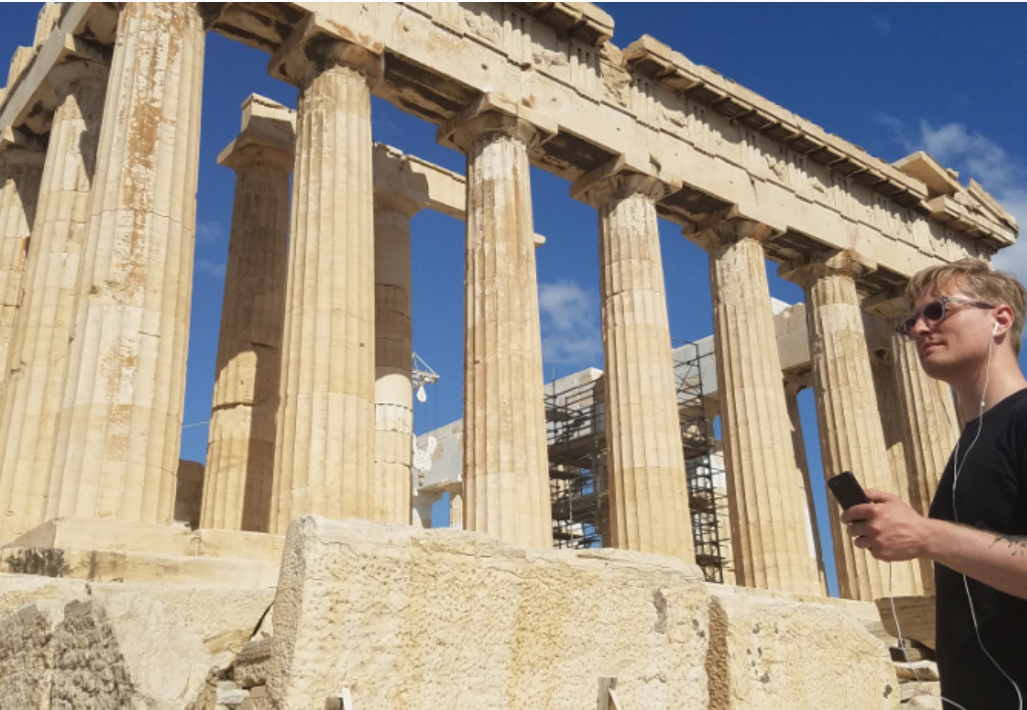 A young man with a cell phone and ear buds standing outside the Acropolis in Athens