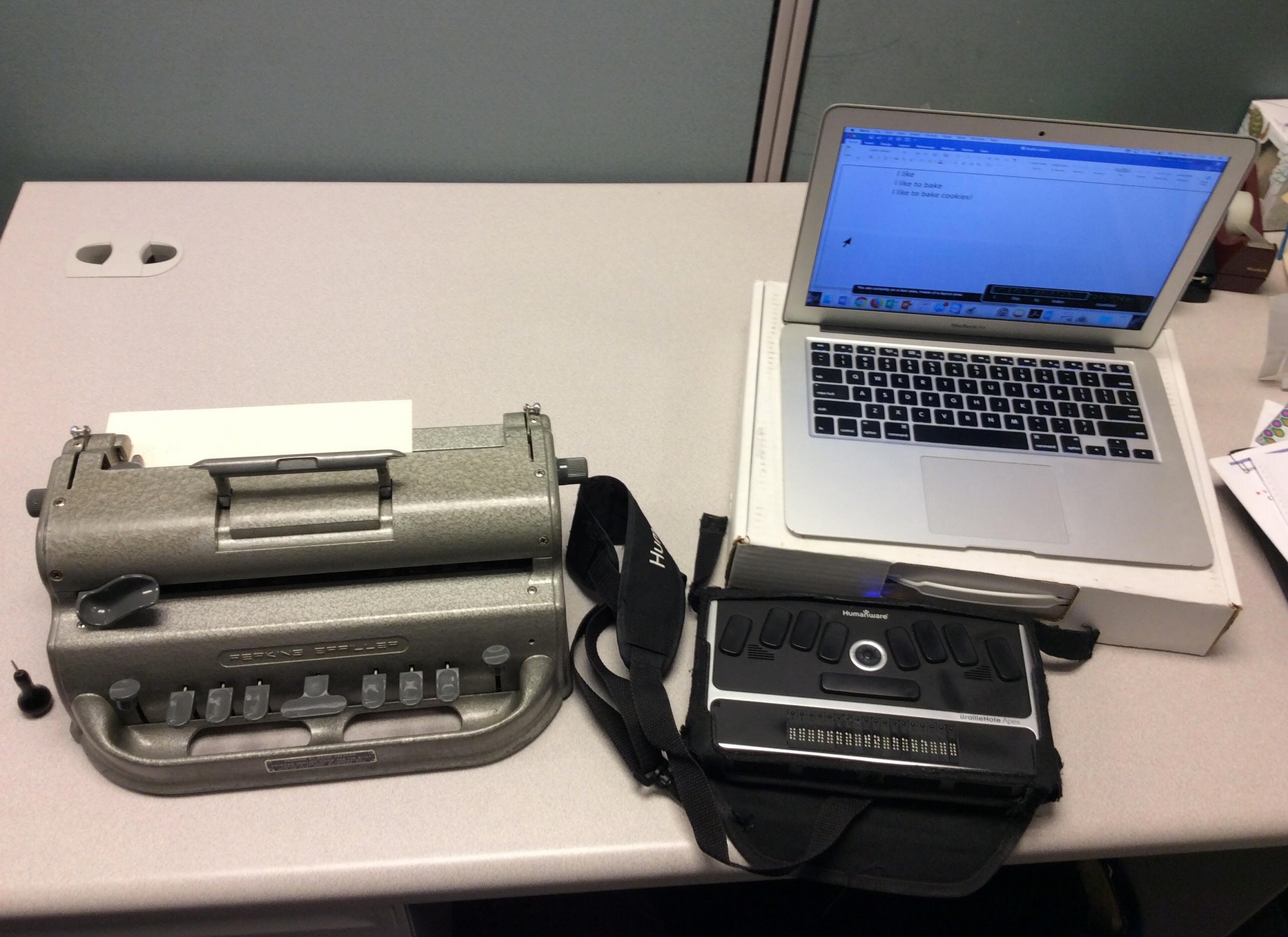  braillewriter, braille eraser, and a braille notetaker paired with a computer.  The braille display reads “I like to bake”.”