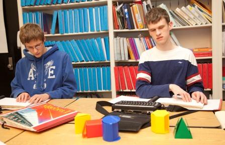 Two boys read braille textbooks with mathematical manipulatives on table.
