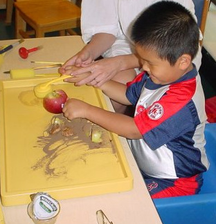 Young boy explores objects on tray, including apple and sand.