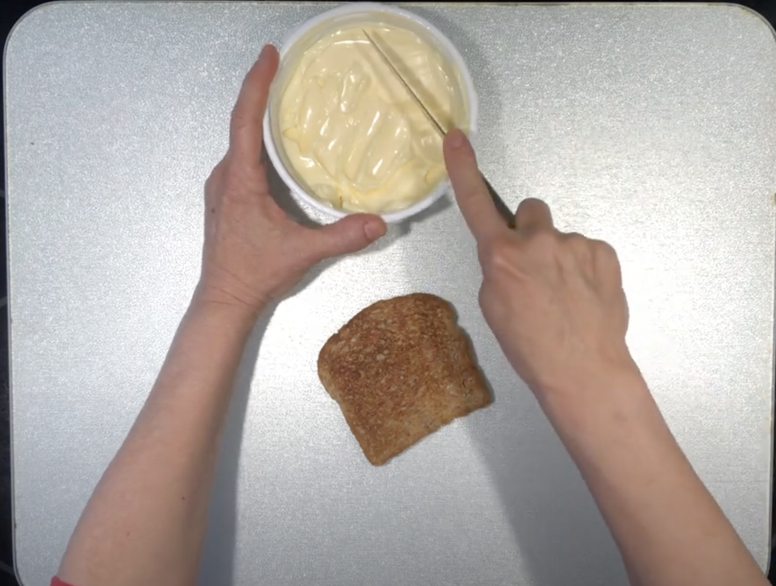 Left hand holding tub of mayonnaise and right hand placing knife in tub with piece of toast in foreground