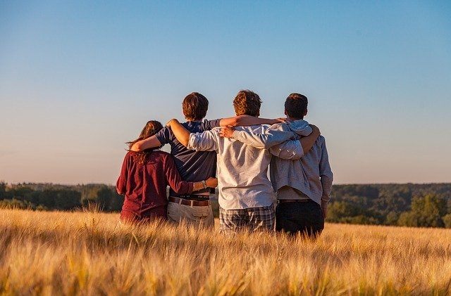 Four teens from behind in a grassy field with arms on eachothers' shoulders